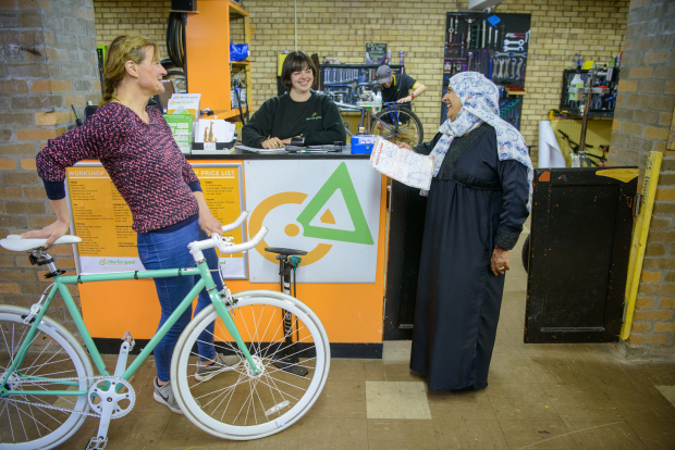 Two women stand, talking at a counter in Bike for Good's shop. A third woman stands behind the counter smiling. The women to the left of the image is holding a green and white bike.