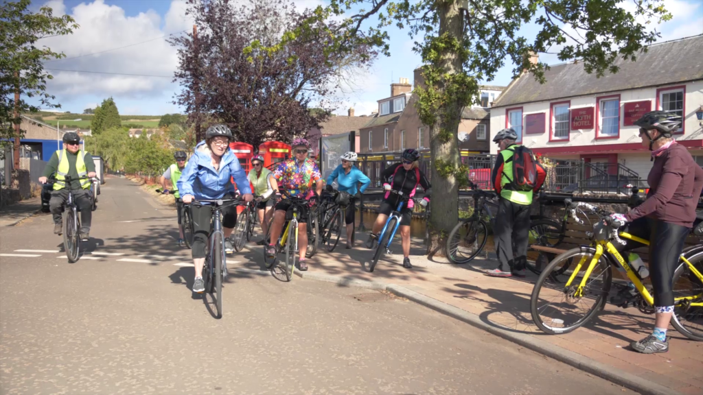A group of people on bikes leaves a village centre. 