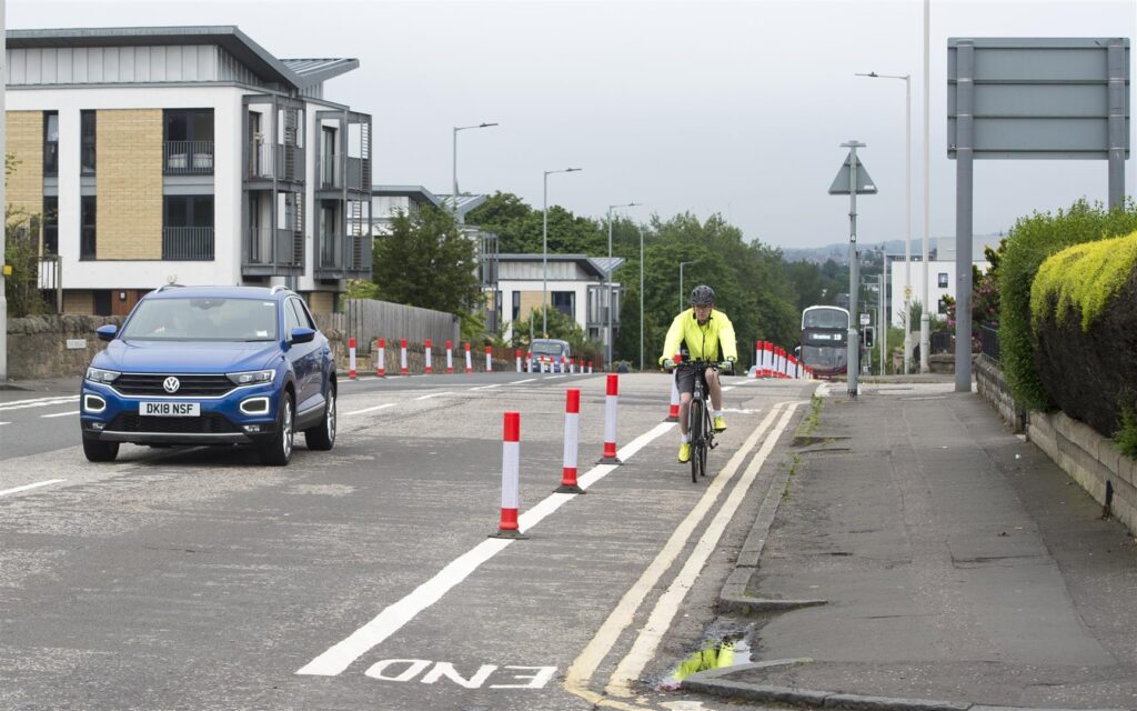 A person cycles uphill, seperated from motor traffic by orange and white stripped cylinders. A blue car is to one side of the cyclist, keeping a safe distance.