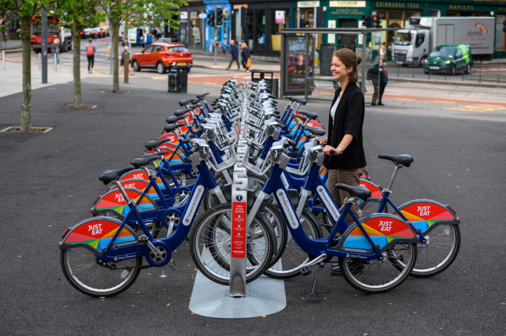 A person unlocks a Transport for Edinburgh hire bike from a rack on Bristo Square. 
