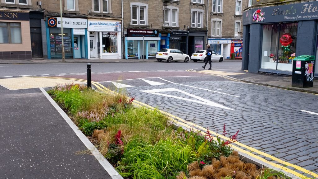 One person is showing crossing the street at the entrance to Balmore Street. Newly installed gardens are shown in the foreground.