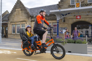 Adult cyclist with child in bike seat cycling on cycle path in front of Stirling Train Station