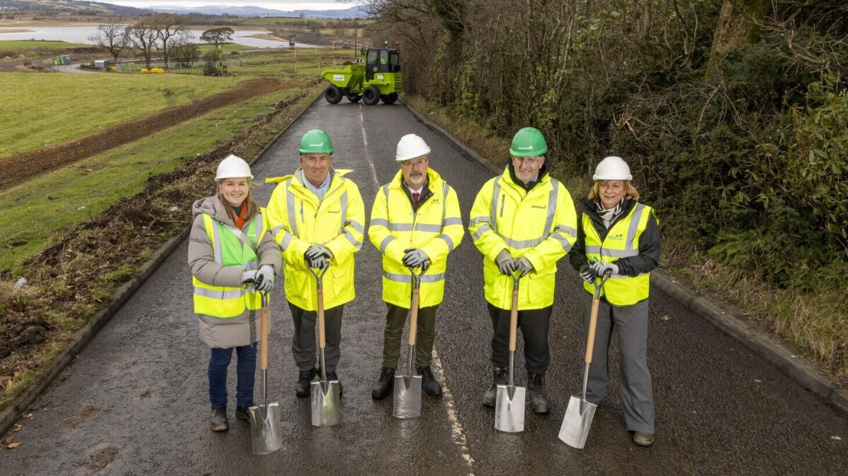 5 adults wearing hi-vis outerwear and protective hard hats stand with a shovels on road