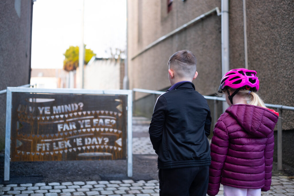 Two children look at a decorative metal barrier featuring phrases in the Caithness dialect. The barrier has replaced two existing barriers in order to improve accessibility for people using wheelchairs, mobility scooters and with buggies.