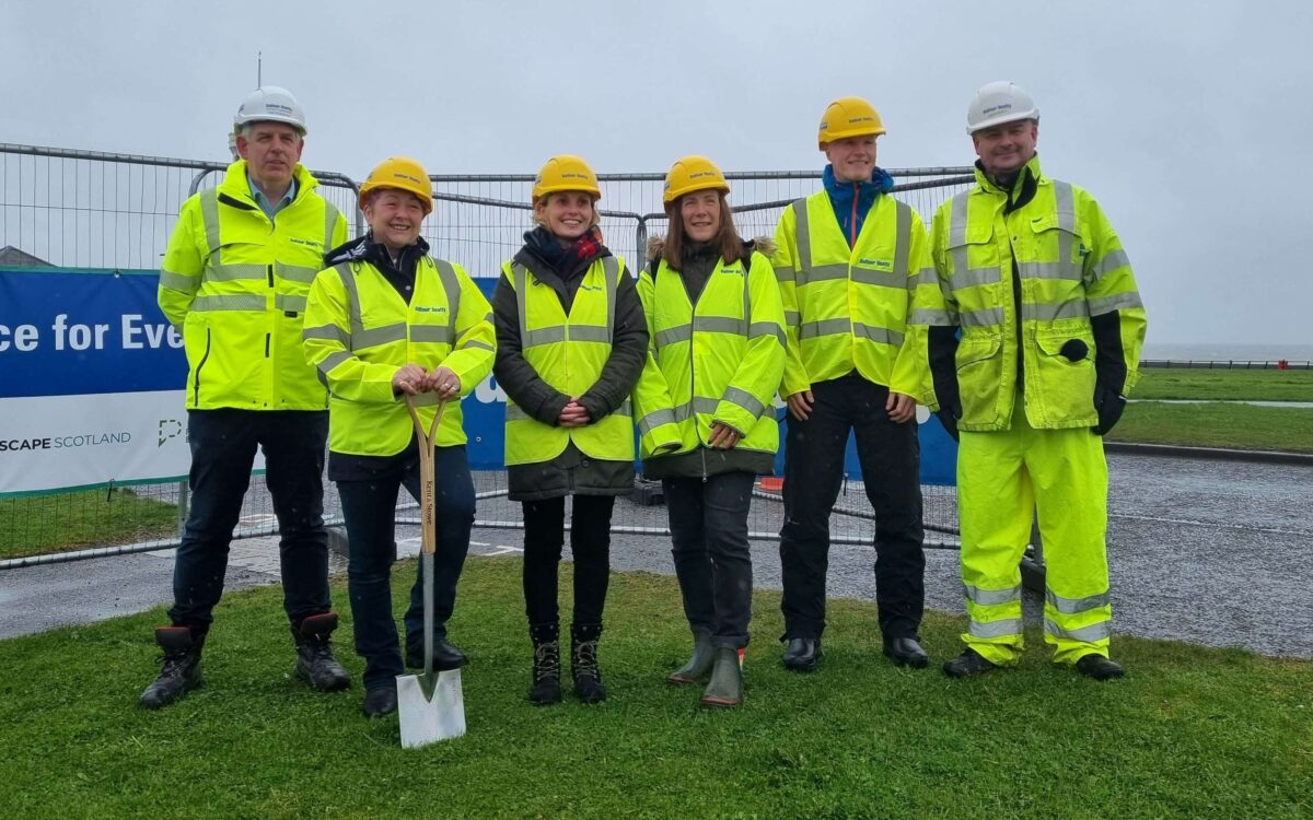 6 adults wearing hi-vis outerwear and protective hard hats stand with a shovel on grass near construction site