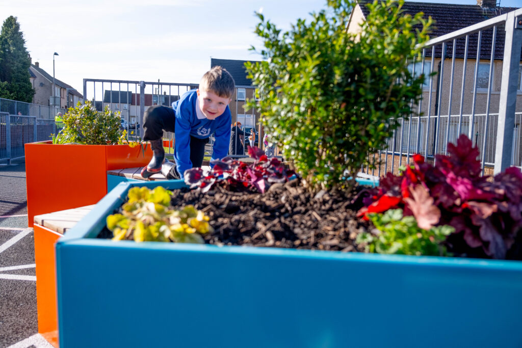 A school pupil is shown playing on a piece of outdoor equipment in the grounds of their primary school. Flowers in a planter are shown in the foreground.