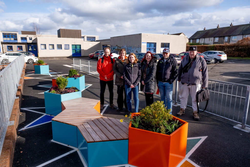 Six people representing the Easter Carmuirs Pocket Places project partners are shown standing by the newly-installed seating area in the grounds of a primary school.